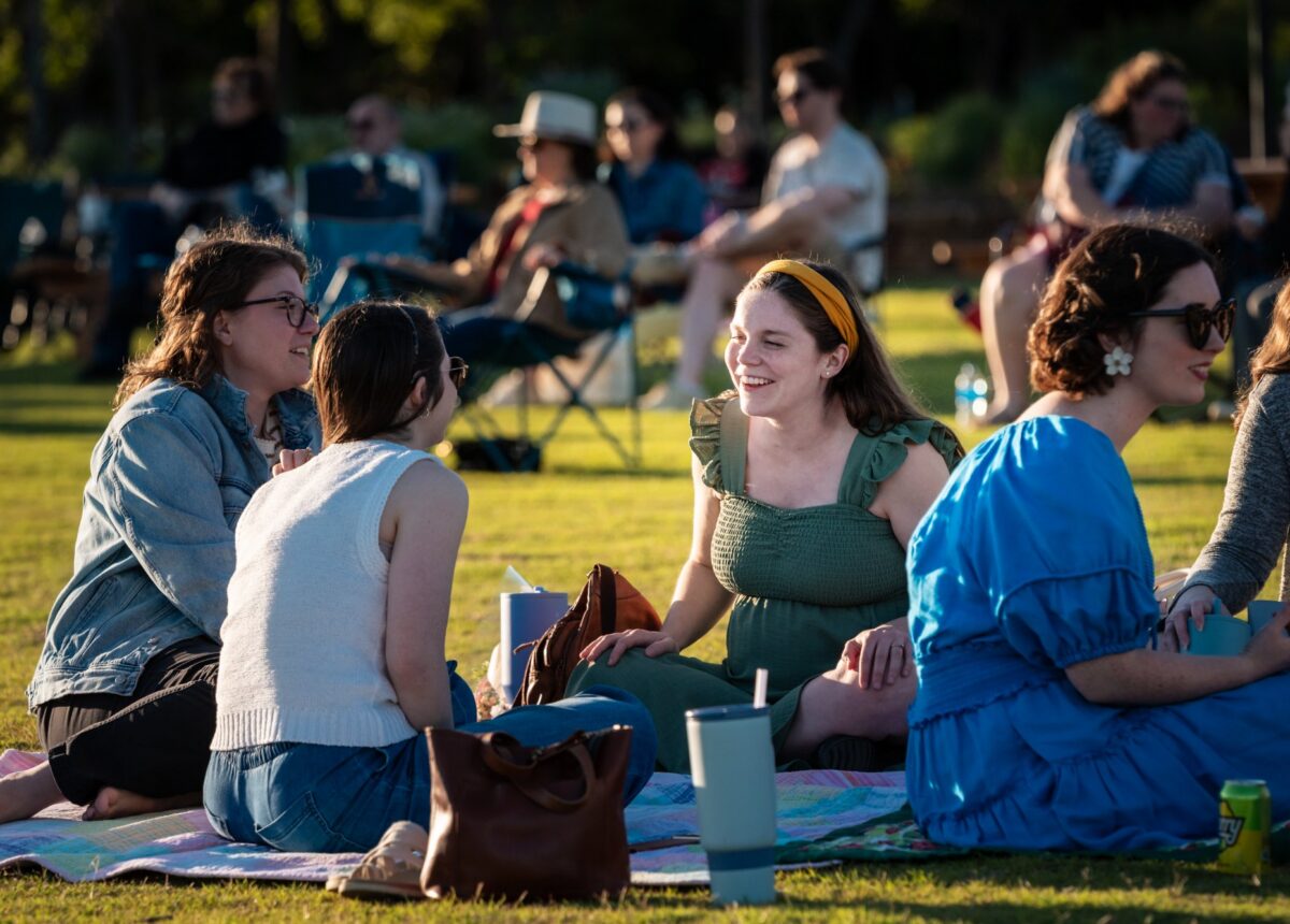 Friends sitting on a picnic blanket on the lawn at Bands & Blooms, with the crowd in lawn chairs behind them in evening light
